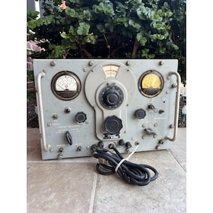 Könnte beinhalten: Ein vintage graues elektronisches Gerät mit mehreren Zifferblättern, Messgeräten und Knöpfen. Das Gerät hat ein zentrales Zifferblatt, zwei kreisförmige Messgeräte und verschiedene Schalter. Ein schwarzes Netzkabel ist unten aufgerollt. Der Text "POWER CIRCUIT SELECTION" ist sichtbar.