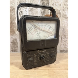 May include: A vintage black Simpson multimeter with a carrying handle. The meter face displays various scales and measurements, including Ohms and Volts. The lower section features dials and switches for different settings. The device is marked "Simpson Electric Co."
