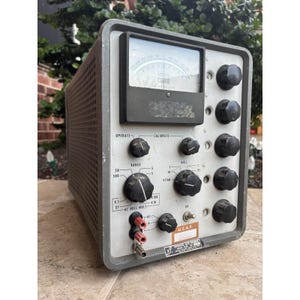 Puede incluir: Dispositivo electrónico gris vintage con múltiples perillas negras, un medidor que muestra "VOLTS" y el texto "U.S.A.F.". El dispositivo tiene forma rectangular con un panel lateral perforado y varias etiquetas de control.