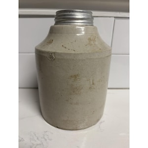 May include: A vintage stoneware jug with a cylindrical body and a slightly flared neck. The jug is a light beige color with some staining and a silver metal screw-on lid. The jug has a rustic, aged appearance.