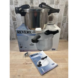 Puede incluir: Una olla a presión Revere de acero inoxidable de 6 litros con asas negras y una caja a juego. La caja presenta una imagen de la olla y el logotipo de Revere. También se incluye un manual y piezas pequeñas.