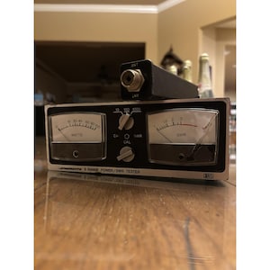 May include: A black and silver 3-range power/SWR tester with two analog meters, a dial, and the text "WATTS" and "SWR". A black antenna connector is on top. The device is on a wooden surface.