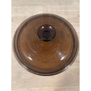 May include: A round, brown glass lid with a dark brown knob in the center. The lid is transparent, allowing a view of the contents below. The lid is designed for cookware.