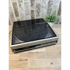Może przedstawiać: Srebrno-czarny gramofon Technics z przezroczystą pokrywą przeciwpyłową. Gramofon ma srebrny panel przedni z czarnymi przyciskami i czarną powierzchnią do odtwarzania płyt. Widoczny jest napis "Technics".