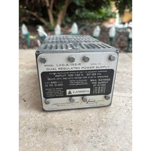 Può includere: Un alimentatore a doppia regolazione, modello LXD-A-152-R, con un involucro grigio metallizzato. Il pannello frontale mostra le specifiche tecniche, tra cui la tensione di ingresso, la frequenza e i valori massimi per alimentazione a 15 VCC. Il logo Lambda è visibile.