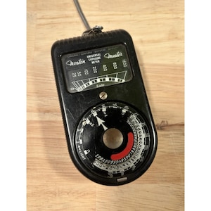 May include: A vintage black Weston Master Universal Exposure Meter. The meter has a dial with numbers and a needle, and a light meter scale. The meter is on a wooden surface.