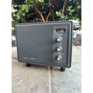 Peut inclure: Une radio vintage grise avec un panneau avant perforé et des boutons de commande. La radio porte l'inscription "YAESU SP-901P" sur le devant. Elle comporte des boutons marqués "TX GAIN", "RX GAIN" et "PATCH". La radio est sur une surface claire.