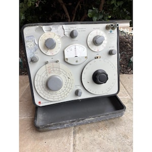 May include: Vintage gray electronic testing equipment with multiple dials, knobs, and gauges. The device has a rectangular shape with a hinged lid and a variety of controls labeled with text. The equipment appears to be used and shows signs of wear.