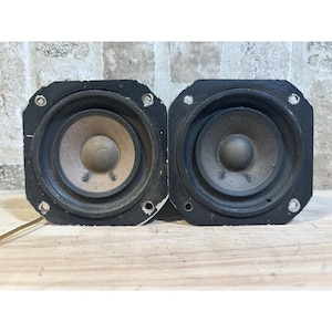 May include: Two vintage black speakers with a square frame and circular speaker cones. The speaker cones are tan and the frames have mounting holes. The speakers are placed side by side on a light-colored wooden surface.