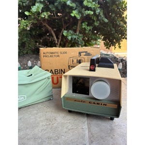 Puede incluir: Proyector de diapositivas automático Cabin U vintage, de color beige y verde. El proyector tiene una lente, un asa negra y la marca "Cabin". También hay una funda de transporte verde y la caja original.