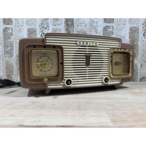 May include: A vintage Zenith radio in brown and cream. It has two clock faces, a speaker grill, and the Zenith logo. The radio includes a power cord. The background is a brick wall.