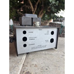 Może przedstawiać: Zabytkowa, srebrna ładowarka terminala Telxon data-Hap z napisami "PHONE", "TERMINAL" i "CHARGER". Ma czarną górę i kilka okrągłych otworów. Widoczny jest również tekst "120 VAC 60 HZ".