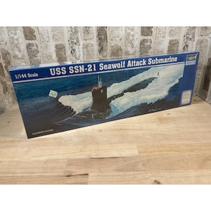 Puede incluir: Caja de modelo del submarino de ataque USS SSN-21 Seawolf. La caja presenta una ilustración del submarino en el mar, con el texto "1/144 Scale" y el nombre del submarino en blanco y azul.