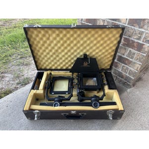 May include: A vintage dark brown photography enlarger in an open suitcase. The enlarger is black with various dials and knobs. The interior of the case is lined with yellow foam. The case has a handle and metal latches.