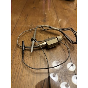 Puede incluir: Una sonda de prueba electrónica con una pinza negra, cables grises y negros y un conector dorado. La sonda está diseñada para el análisis y la medición de circuitos eléctricos. Los cables están dispuestos sobre una superficie de madera.