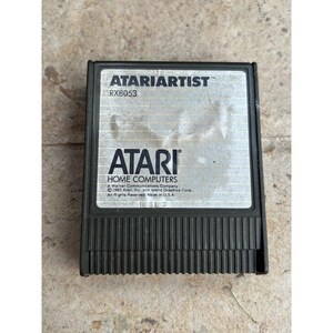 May include: A black Atari cartridge labeled "ATARIARTIST" and "RX8053". The cartridge also features the Atari Home Computers logo and copyright information. The bottom of the cartridge has a series of vertical ridges.