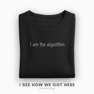 May include: A black crew neck t-shirt featuring the white text I am the algorithm printed on the chest. The garment is folded neatly, displaying a simple round neckline and short sleeves, with the fabric texture visible across the surface.