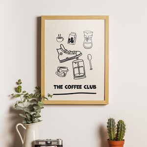 Puede incluir: Un estampado enmarcado con fondo beige presenta dibujos lineales negros de elementos relacionados con el café, incluyendo una zapatilla deportiva, una taza de café, un cartón de leche y una prensa francesa. El texto "THE COFFEE CLUB" está impreso en negro.