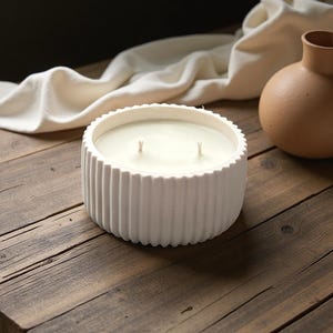 Op de afbeelding: Een witte, ronde kaars met twee lonten staat op een houten tafel. De kaars zit in een geribbelde houder. Een beige vaas en een witte doek staan op de achtergrond.