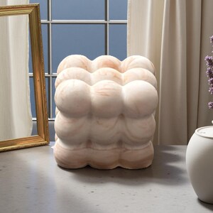 Peut inclure: Un objet décoratif en forme de cube avec un design en forme de nuage. L'objet est de couleur rose clair avec des veines roses plus foncées. Il repose sur une surface grise près d'une fenêtre et d'un miroir doré.