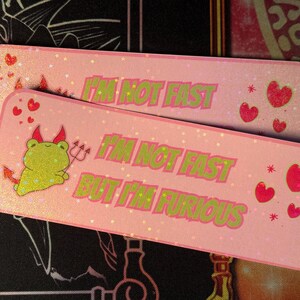 May include: Two pink rectangular stickers with glitter. One sticker features a cartoon frog with devil horns and a trident. The text reads "I'M NOT FAST BUT I'M FURIOUS". Red hearts and stars are scattered across the stickers.
