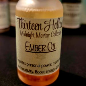 Può includere: Una bottiglia di Ember Oil della collezione Thirteen Hollow Midnight Mortar. L'etichetta è verde chiaro con testo nero che dice "EMBER OIL". L'olio è di colore dorato e il fondo della bottiglia ha una tonalità arancione.