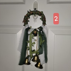 May include: A decorative wreath with a circular vine base, adorned with green and white fabric, faux leaves, and three small brass bells. A small bottle is attached to the center. The wreath is hanging on a white door.