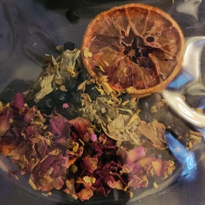 Può includere: Un sacchetto di plastica trasparente pieno di erbe e piante essiccate. Il mix include fette d'arancia essiccate, petali di rosa e altri ingredienti verdi e blu scuro. Il contenuto è un mix di consistenze e colori, che crea un'estetica naturale e organica.