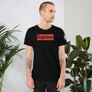 May include: Black t-shirt with the word "Suspicious" in a red rectangular box on the front. The shirt has short sleeves and a crew neck. A small square graphic is visible on the right sleeve.