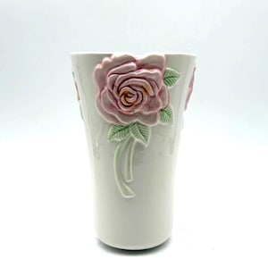 May include: A white ceramic vase with a raised pink rose design. The rose has detailed petals and green leaves. The vase is cylindrical and has a smooth, glossy finish. A decorative floral accent for the home.