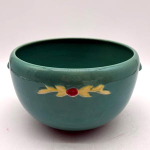 May include: A teal ceramic bowl with a smooth, glossy finish. The exterior features a centered decorative motif consisting of two yellow, leaf-like shapes flanking a small, raised red circular element. The bowl has a wide, open top.