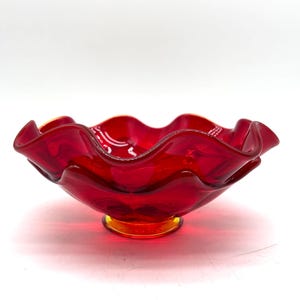 May include: A vibrant red glass bowl featuring a wide, ruffled, and undulating rim. The bowl is supported by a small, circular base that transitions into a subtle amber-orange hue at the very bottom, contrasting with the deep red glass body.