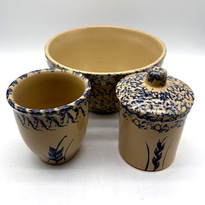 May include: A set of three ceramic kitchen items featuring a tan base color with blue sponge-painted rims and blue wheat stalk motifs. The set includes a large open bowl, a smaller open cup, and a lidded canister with a matching blue knob.