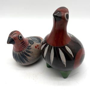 May include: Two hand-painted ceramic bird figurines. The smaller bird features a light beige body with red, black, and white patterns. The larger bird is taller, with a dark brown and red body, white feather-like markings, and three green feet.