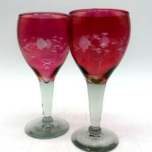 May include: Two stemmed wine glasses with ruby red bowls and clear stems and bases. Each glass features an etched floral design. The glasses are set against a white background.