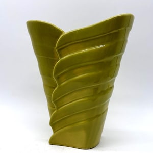 May include: A chartreuse-colored ceramic vase with a unique, sculptural design. The vase features a flared, asymmetrical shape with a series of curved, ridged details that wrap around the body. The vase has a glossy finish and a rectangular base.