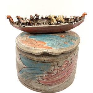 May include: A ceramic jar with a lid, featuring a hand-painted design in blue and pink. A small, boat-shaped sculpture with animal figures sits atop the jar. The sculpture and jar are in earthy tones.