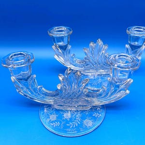 May include: A clear glass candelabra with four candle holders. The candelabra has a floral design on the base and decorative leaf-like arms. The background is a solid blue.