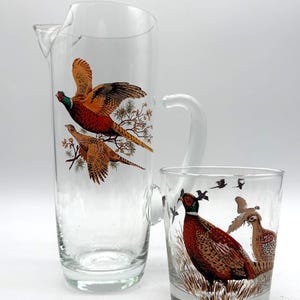 May include: A clear glass pitcher and tumbler set. The pitcher has a handle and a spout. Both pieces are decorated with images of pheasants in flight and on the ground, featuring brown, orange, and green colors. The set is on a white background.