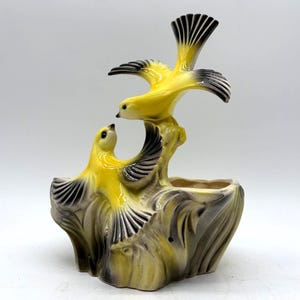 May include: A ceramic planter featuring two yellow birds with black and white wings perched on a tree stump. The birds are facing each other, with the top bird's wings spread. The planter has a neutral color palette with a glossy finish.