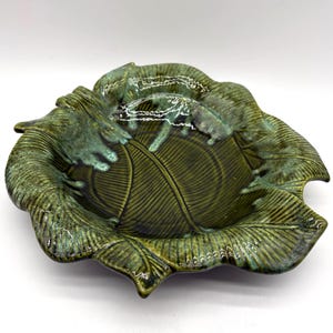 May include: A green ceramic leaf-shaped dish with a glossy finish. The dish features detailed veining and a textured surface, with a darker green interior and lighter green edges. This decorative piece is ideal for serving or display.