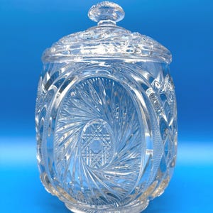 May include: A clear, cut-glass jar with a lid. The jar has an oval design on the front and is decorated with intricate patterns. The lid has a small, round knob. The jar is on a blue background.