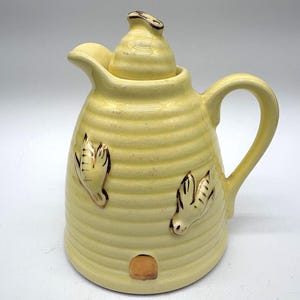 May include: A yellow ceramic creamer with a lid and handle. The creamer has a beehive design with embossed bee accents and a small opening. The creamer is a light yellow colour with gold accents.