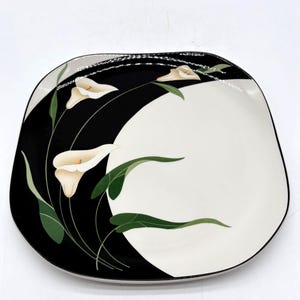 May include: A square ceramic serving plate with a black and white floral design. The plate features a black curved section with cream-colored calla lilies and green leaves. The other half of the plate is white.