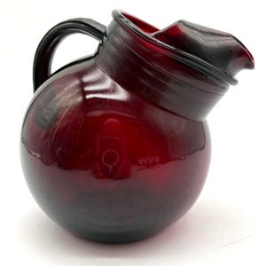 May include: A ruby red glass pitcher with a rounded body and a curved spout. The pitcher has a black handle and a dark rim. The glass is transparent, allowing light to pass through. This vintage-style pitcher is suitable for serving beverages.