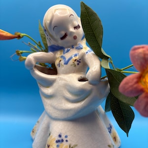 May include: A vintage ceramic figurine of a girl in a dress, with closed eyes and a serene expression. The dress is white with yellow and blue floral accents. The figurine is holding a basket, and is decorated with a blue scarf. The background is a solid blue.