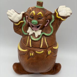 May include: Brown ceramic clown cookie jar with a smiling face, white gloves, and green and yellow accents. The jar has a lid and is in the shape of a clown with arms raised. A vintage kitchen collectible.