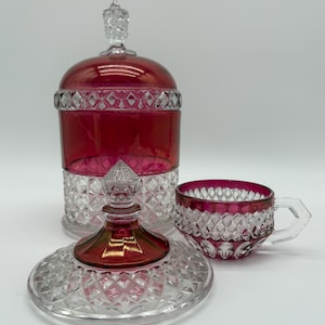 May include: A ruby red and clear glass tea set with diamond-cut detailing. The set includes a lidded jar, a saucer, and a teacup with a handle. The glass has a vintage aesthetic.