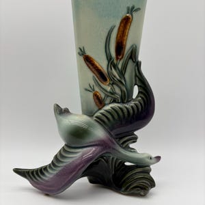 May include: A ceramic vase with a light blue background and a design of cattails. The vase is supported by a sculpture of two birds with green and purple feathers. The vase is approximately 8 inches tall.