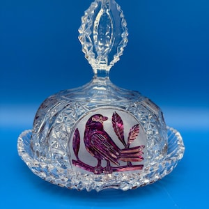 May include: A clear glass butter dish with a decorative bird design. The lid features a faceted handle and a circular design with a purple bird perched on a branch. The dish has a scalloped edge and a diamond pattern.